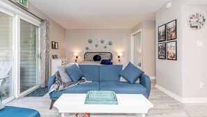 Un salon moderne avec un canapé bleu, une table basse blanche et un lit avec une tête de lit. On y trouve des lampes fixées au mur, une horloge et des tableaux encadrés.