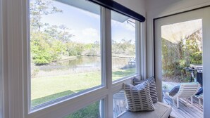 A room with large windows overlooking a pond and greenery, a deck with outdoor furniture, and a built-in bench with cushions. - Lakefront home w/ Private Dock + Golf Cart l 8 min. to beach (St. Simons Island)
