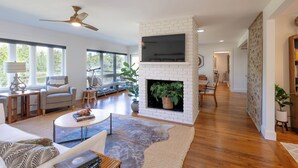 A modern living room with a fireplace, large windows, and a ceiling fan. - Lakefront home w/ Private Dock + Golf Cart l 8 min. to beach (St. Simons Island)