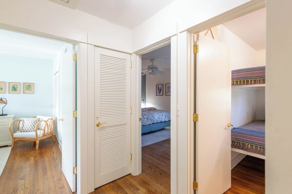 A room with a wooden floor, a bed with a blue bedspread, a white door with a louvered panel, and a small chair with a patterned cushion. - Lakefront home w/ Private Dock + Golf Cart l 8 min. to beach (St. Simons Island)