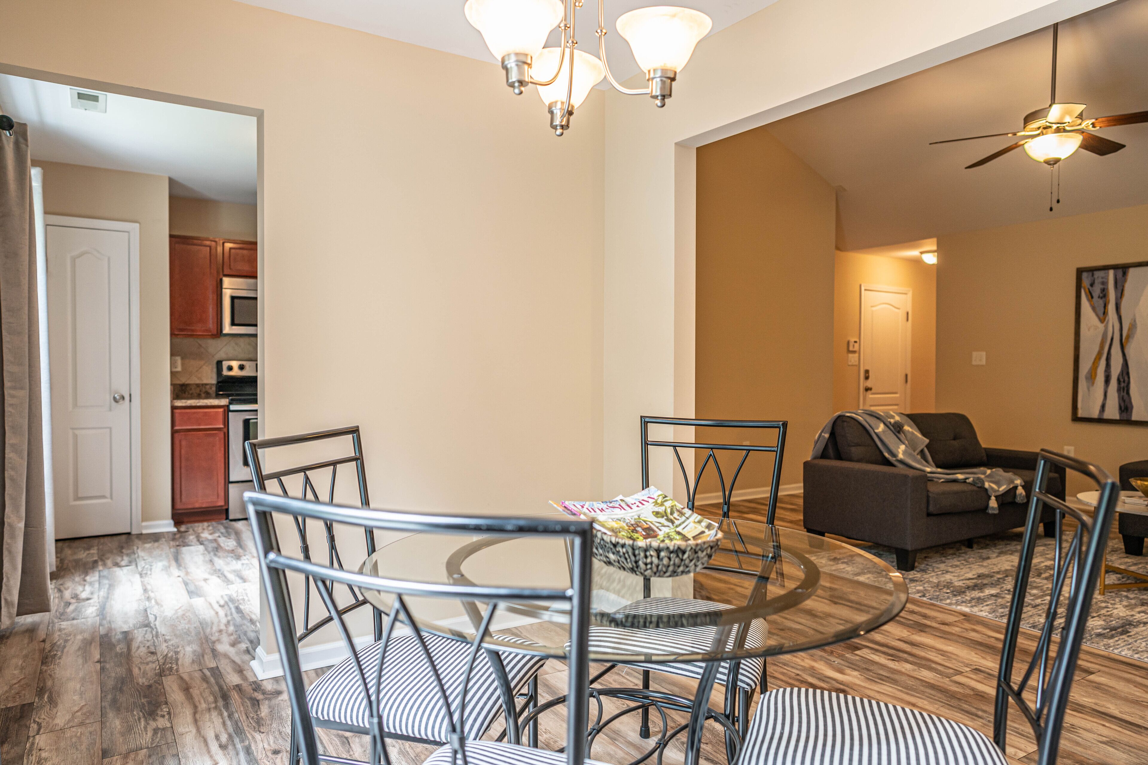 A modern living room with a dining area, featuring a round glass table, metal chairs, and a ceiling fan.