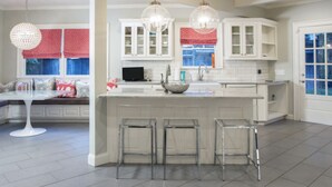 A modern kitchen with white cabinetry, a central island with stools, pendant lighting, and a view of the outside through a window with red curtains. - Beautiful Bright 6 bedroom home in the heart of Buckhead (Atlanta)
