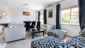 A modern living room with a dining area, featuring a fireplace, a sofa set, a coffee table, and a large window with a view of buildings. - Cantinho da fraternidade - 3 bed apartment (Portimão)