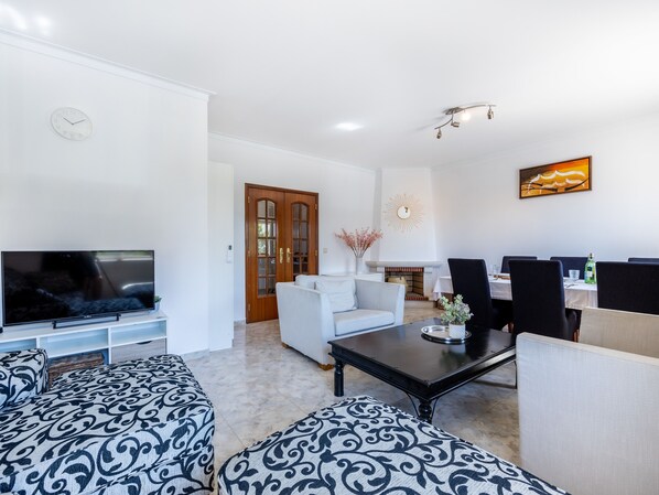 A modern living room with a flat-screen TV, patterned sofas, a dining area with chairs, and a fireplace. - Cantinho da fraternidade - 3 bed apartment (Portimão)