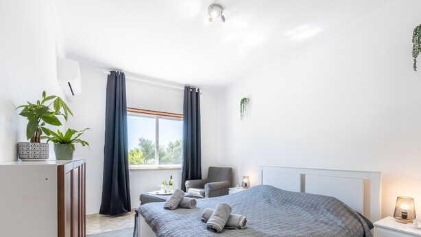 A modern bedroom with a large bed, a desk, a chair, a window with curtains, and a potted plant. - Cantinho da fraternidade - 3 bed apartment (Portimão)