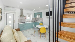 A modern kitchen with teal cabinets, a white countertop, and a built-in stove. There is a dining area with a white table and two chairs, one yellow and one gray. A wooden staircase is visible on the right. - House (Porto)