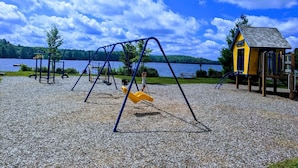 A playground with swings and a slide, a small yellow playhouse, and a lake in the background.