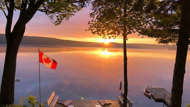 A lakeside deck with a Canadian flag, outdoor seating, and a dock extending into the water.