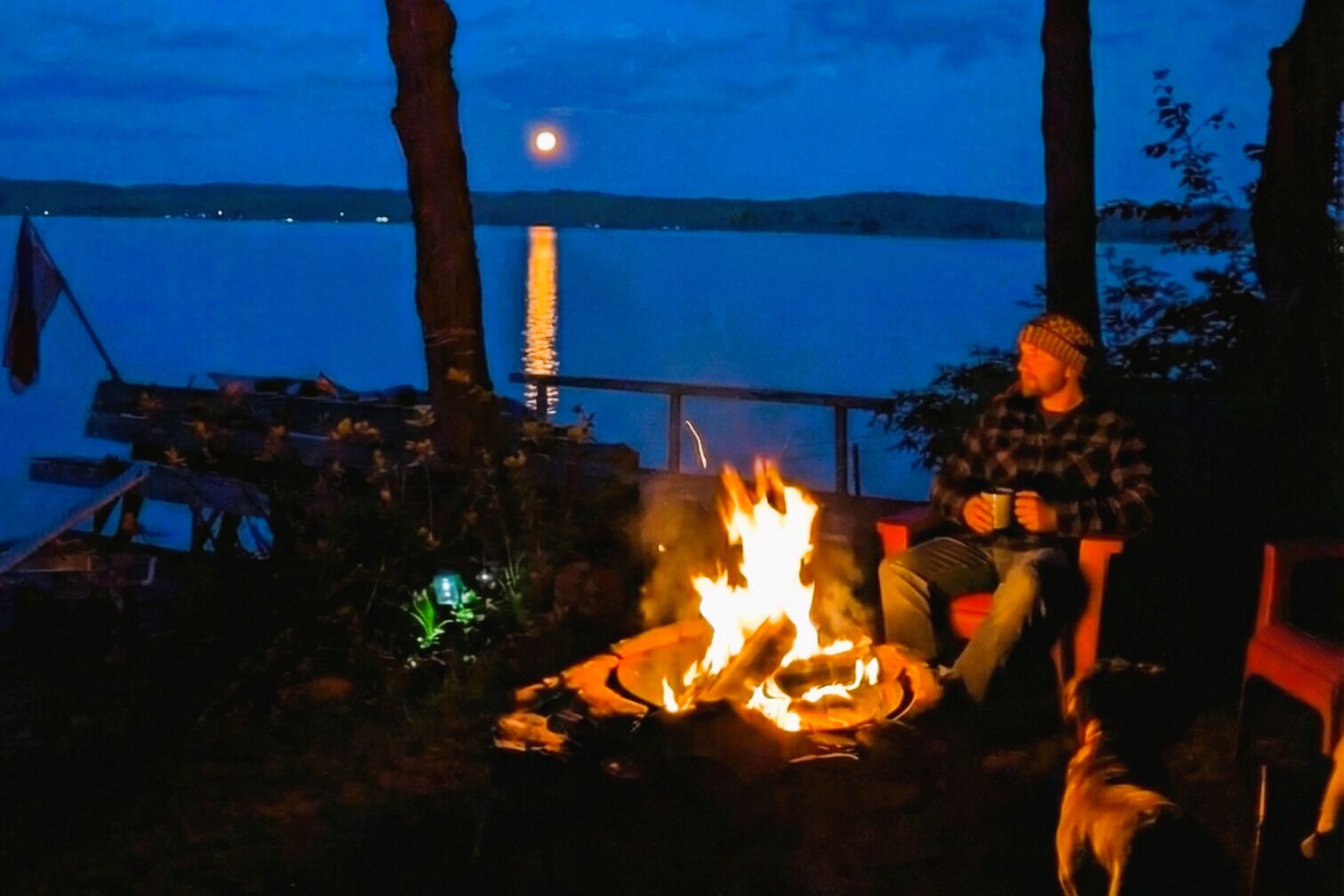 A person sitting by a campfire with a lake and a moon in the background.