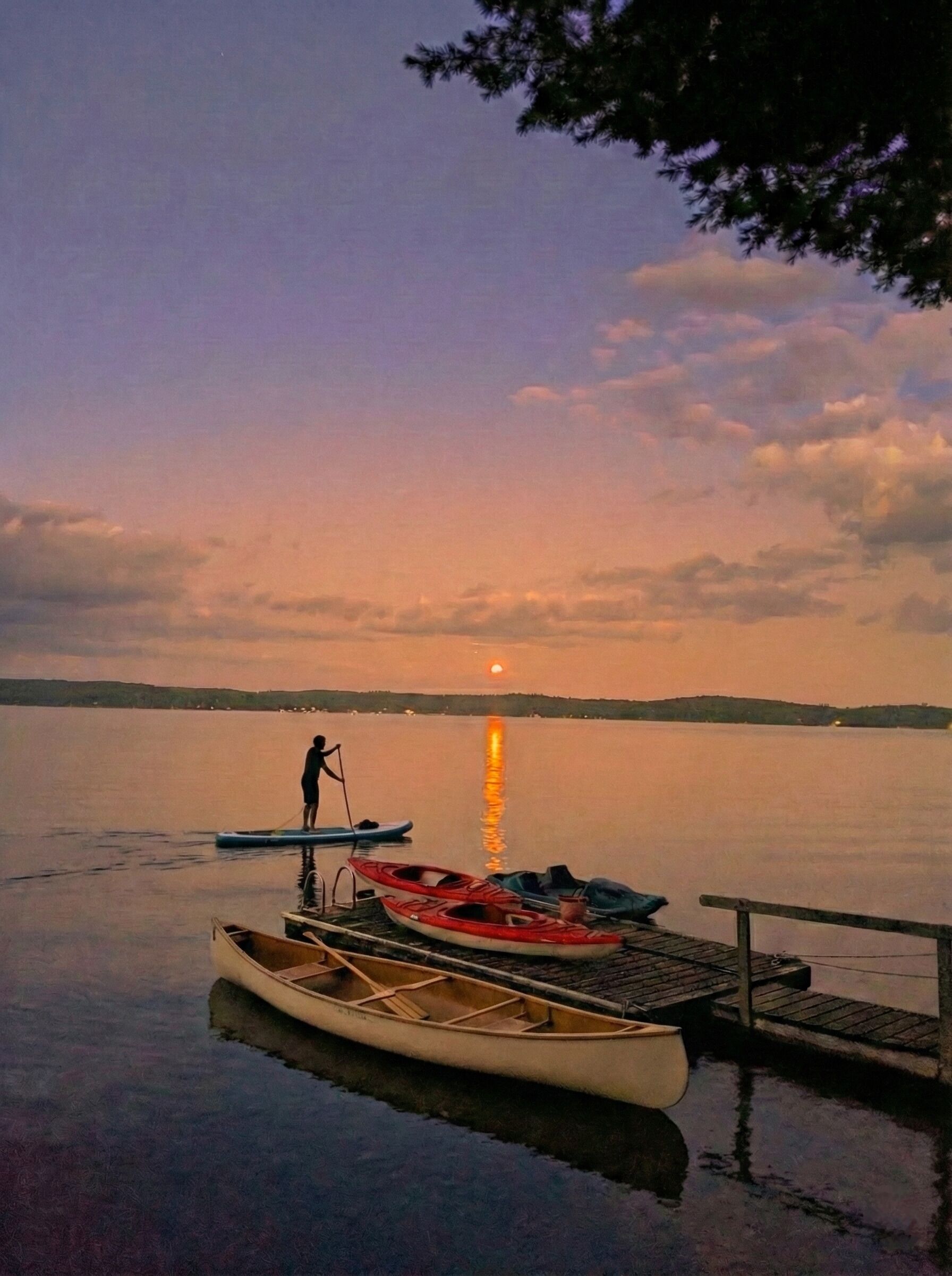 A calm lake at sunset with a person paddling a canoe, docked boats, and a wooden pier.