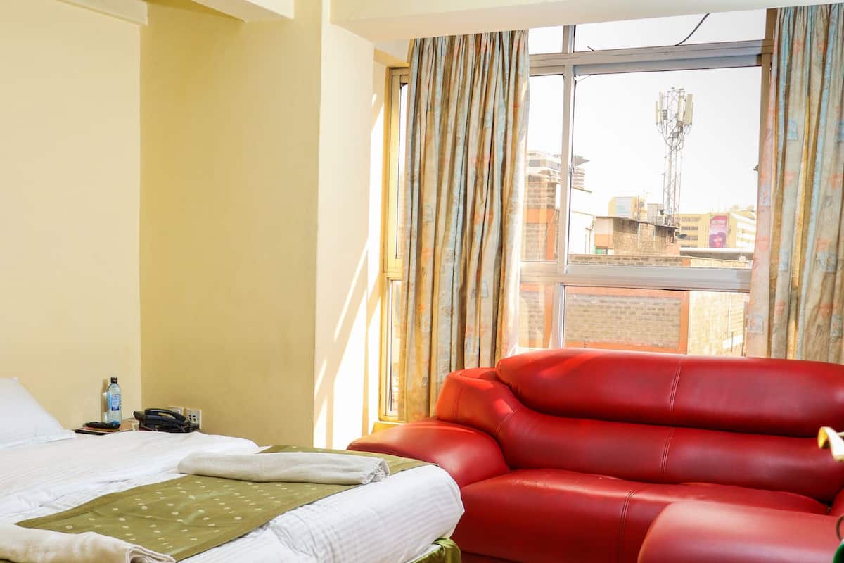 A hotel room with a bed, a red sofa, a window with curtains, and a view of buildings outside.