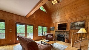 A wooden cabin interior with a sofa, armchair, fireplace, TV, and large windows. - Yonah View Lodge - 2 BR Cabin w/ Spacious Decks, Hot Tub, Fire Pit, Near Helen (Cleveland)
