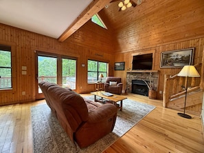 A wooden cabin interior with a sofa, armchair, fireplace, TV, and large windows.