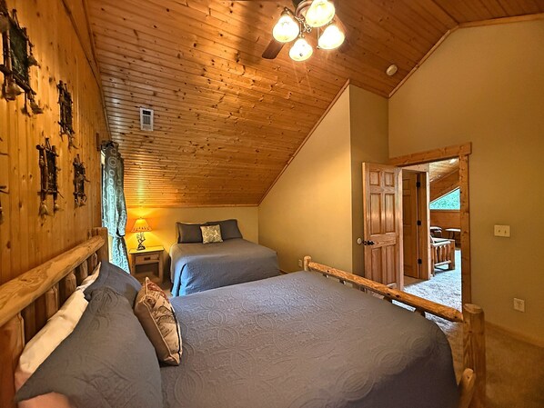 Een kamer met houten balken in het plafond, twee bedden met grijze beddengoed, een houten deur die leidt naar een balkon, en een klein tafeltje met een lamp.