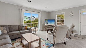 A living room with a sofa, coffee table, and armchair, a TV on a stand, and a view of a beach through large windows. - Secret Spot: Steps to Beach, Dog-Friendly Duck (Duck)
