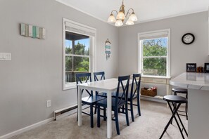 A dining area with a white table and blue chairs, large windows with views of greenery, a chandelier, and a clock on the wall.