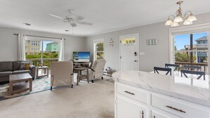 A spacious living area with a ceiling fan, a flat-screen TV, a dining table with chairs, and a large window offering a view of the beach and buildings. - Secret Spot: Steps to Beach, Dog-Friendly Duck (Duck)