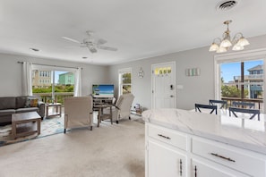 A spacious living area with a ceiling fan, a flat-screen TV, a dining table with chairs, and a large window offering a view of the beach and buildings.