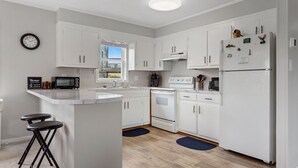 A modern kitchen with white cabinetry, a central island with stools, a built-in oven, a refrigerator, and a clock on the wall. - Secret Spot: Steps to Beach, Dog-Friendly Duck (Duck)