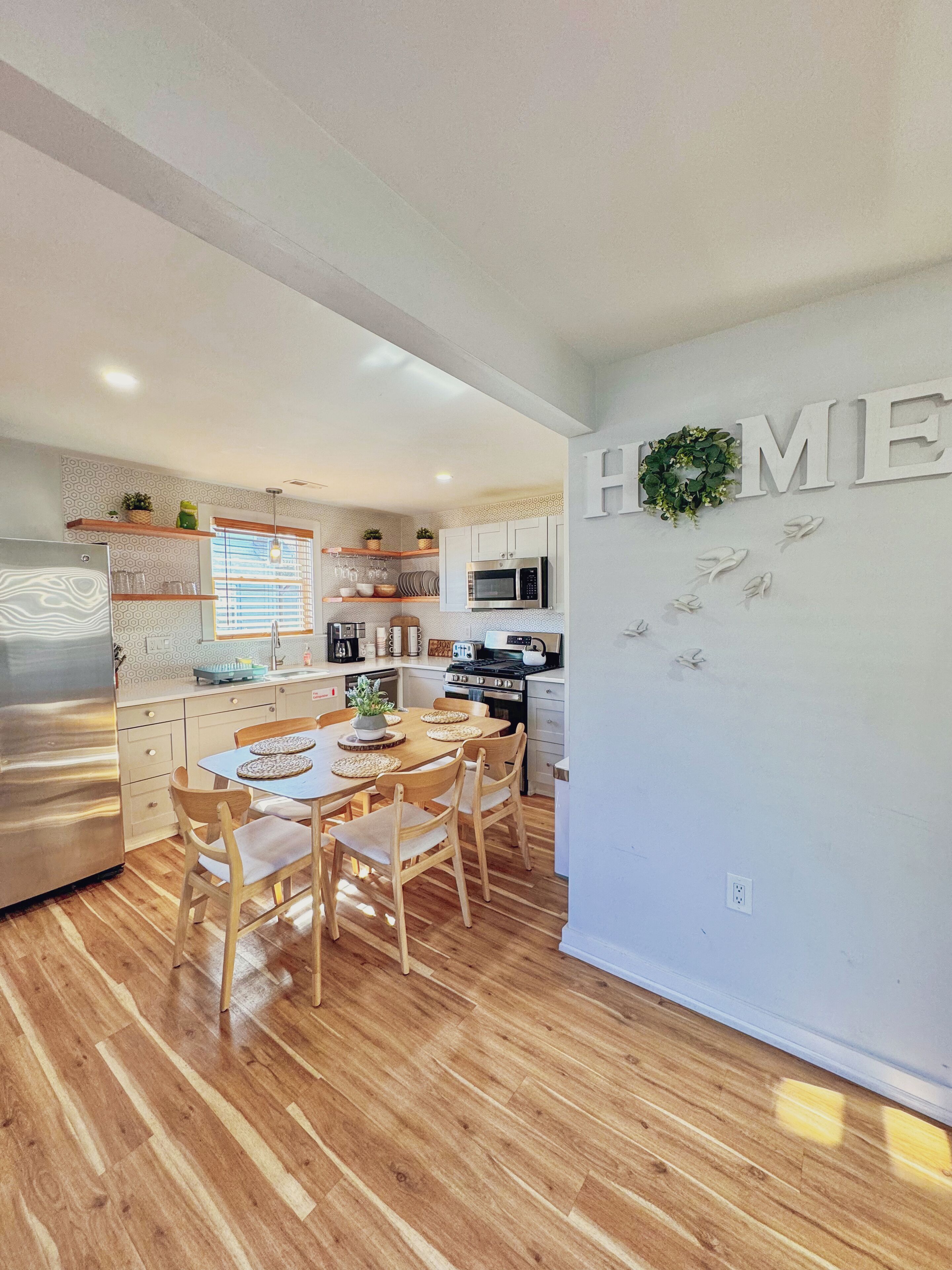 A modern kitchen with white cabinets, a dining table with chairs, and a 'HOME' sign on the wall.