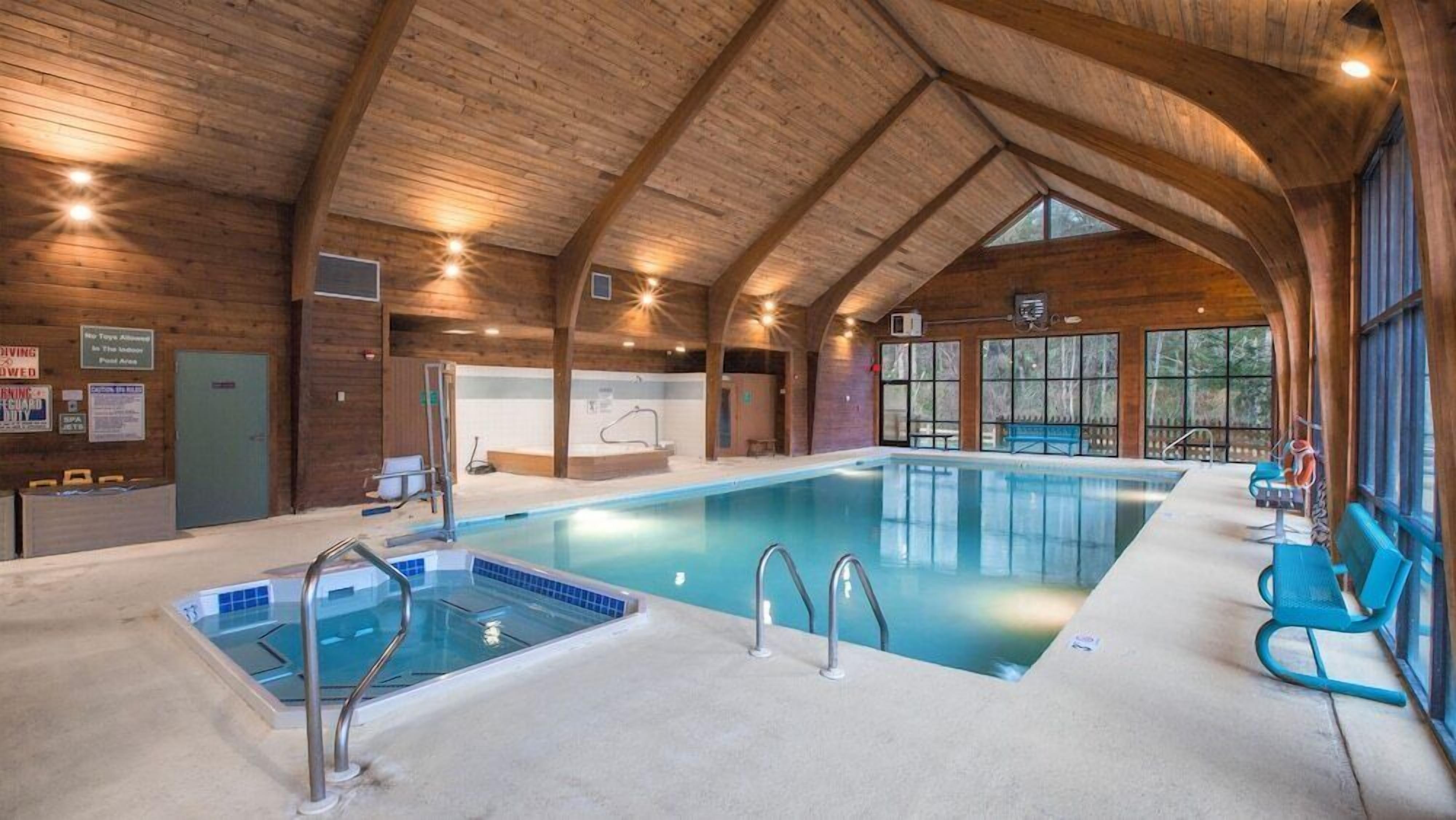An indoor swimming pool with wooden beams, a changing room, and a view of trees through large windows.