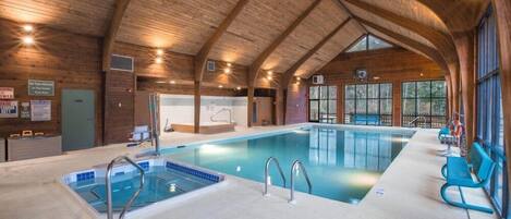 An indoor swimming pool with wooden beams, a changing room, and a view of trees through large windows.