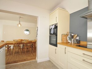 A kitchen with wooden cabinets, a stainless steel range hood, and a dining area with a wooden table and chairs. - The Porthole (Port Isaac)