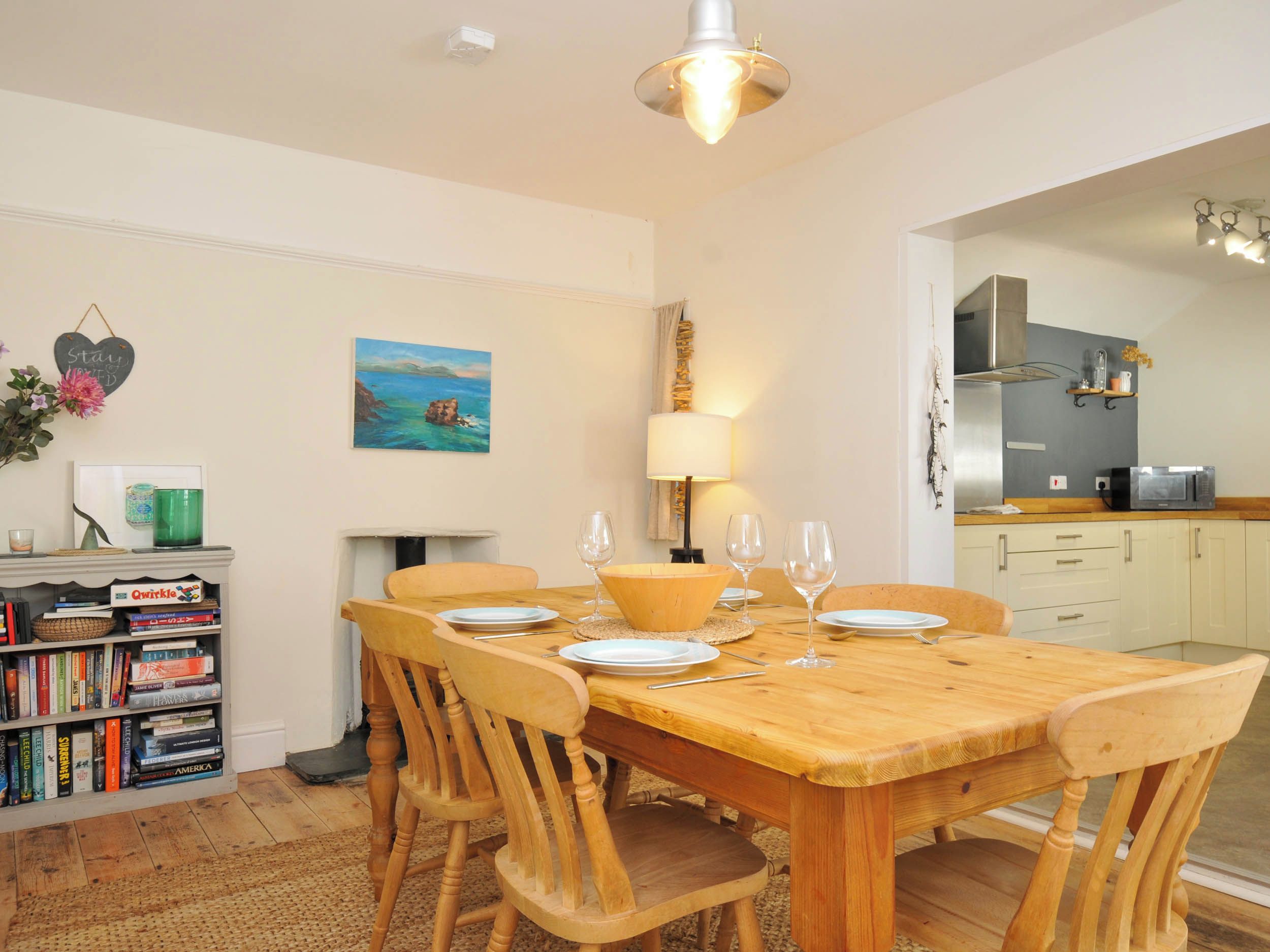A dining area with a wooden table and chairs, a bookshelf with books, a painting, and a kitchen area with a microwave.