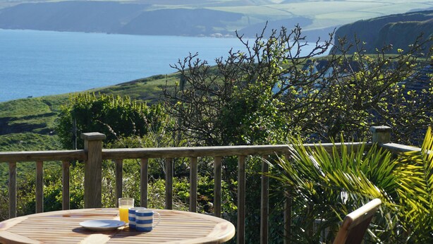 Une terrasse en bois avec une table dressée pour le petit-déjeuner, offrant une vue sur un paysage côtier avec des collines et la mer.