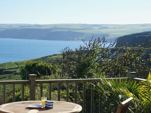 Un terrazzo in legno con un tavolo apparecchiato per la colazione, con vista su un paesaggio costiero collinare e il mare.
