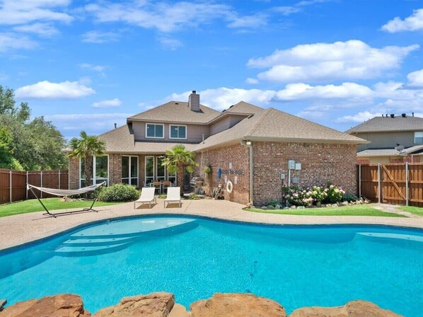 A residential backyard with a swimming pool, a brick house, and a hammock.