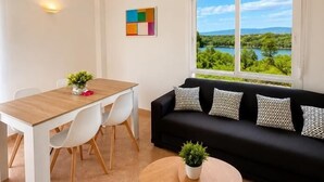 A room with a dining table, white chairs, a black sofa with patterned cushions, a view of a lake, and a colorful abstract painting on the wall. - APARTBEACH LOS JUNCOS 1 RESIDENTIAL (La Pineda)