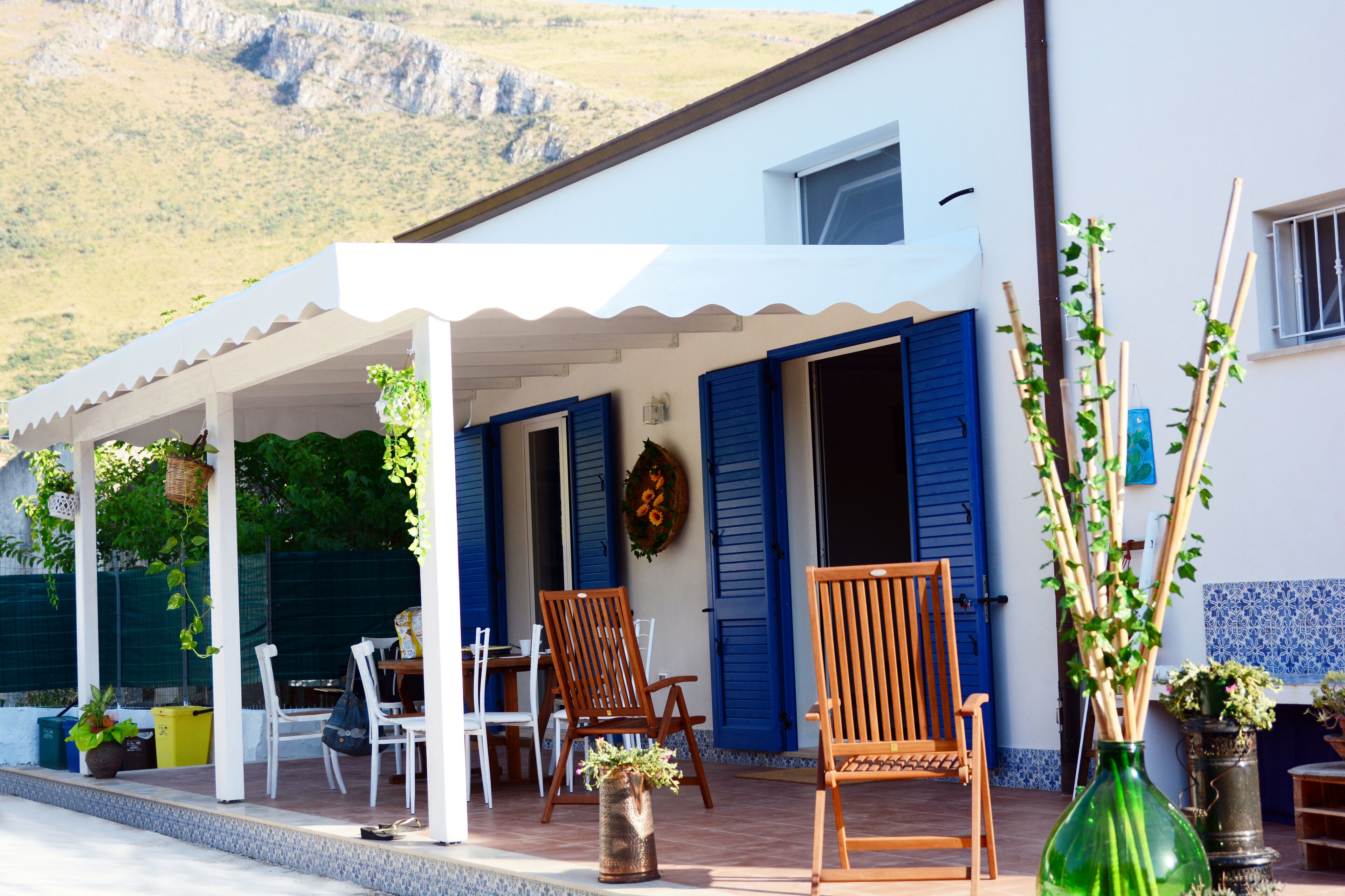 A covered outdoor seating area with chairs and tables, a green vase with plants, and a building with blue shutters.