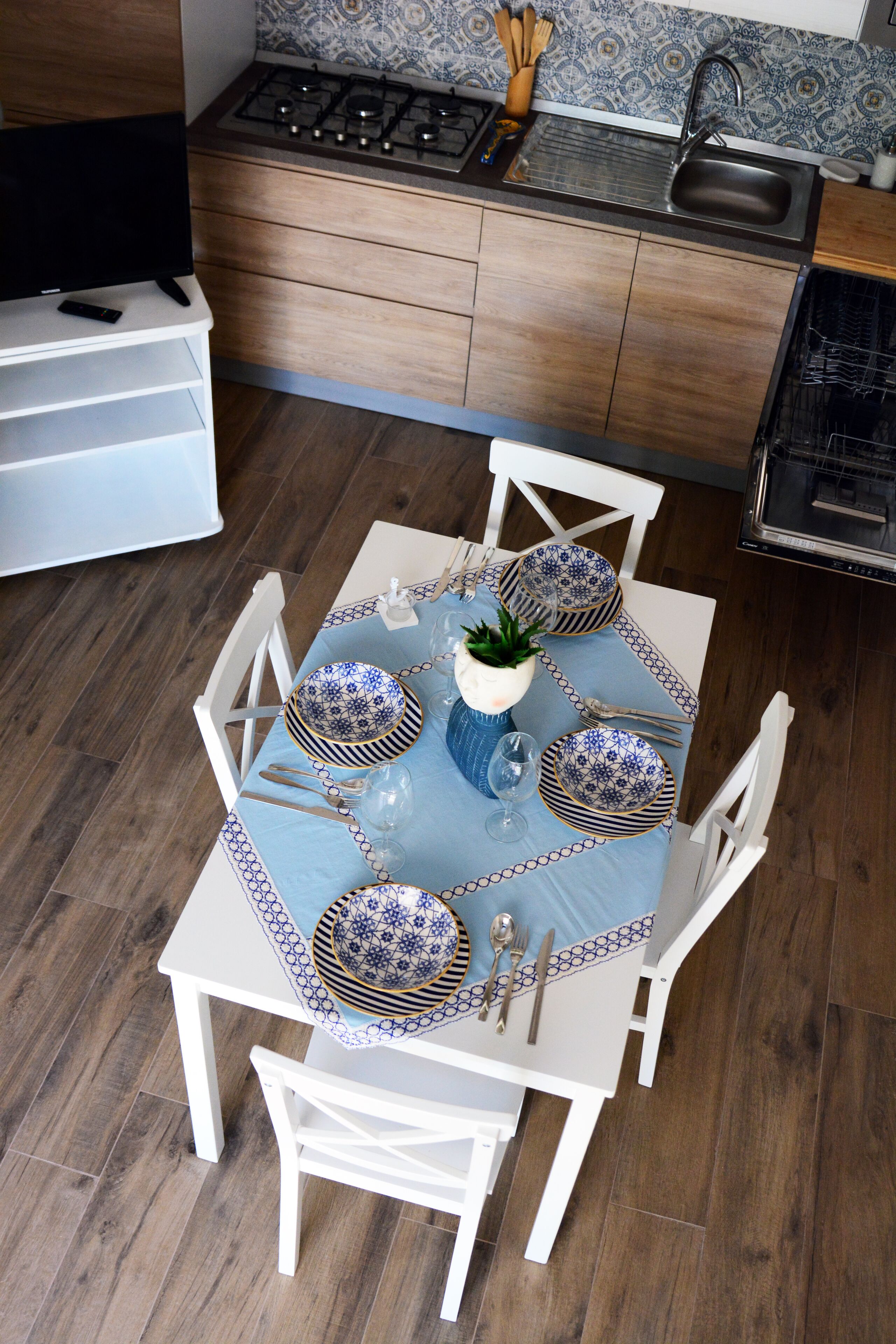 Un coin repas avec une table carrée dressée pour quatre personnes, entourée de chaises blanches. Une petite plante en pot est posée sur la table, et on y trouve des assiettes, des couverts et des verres.