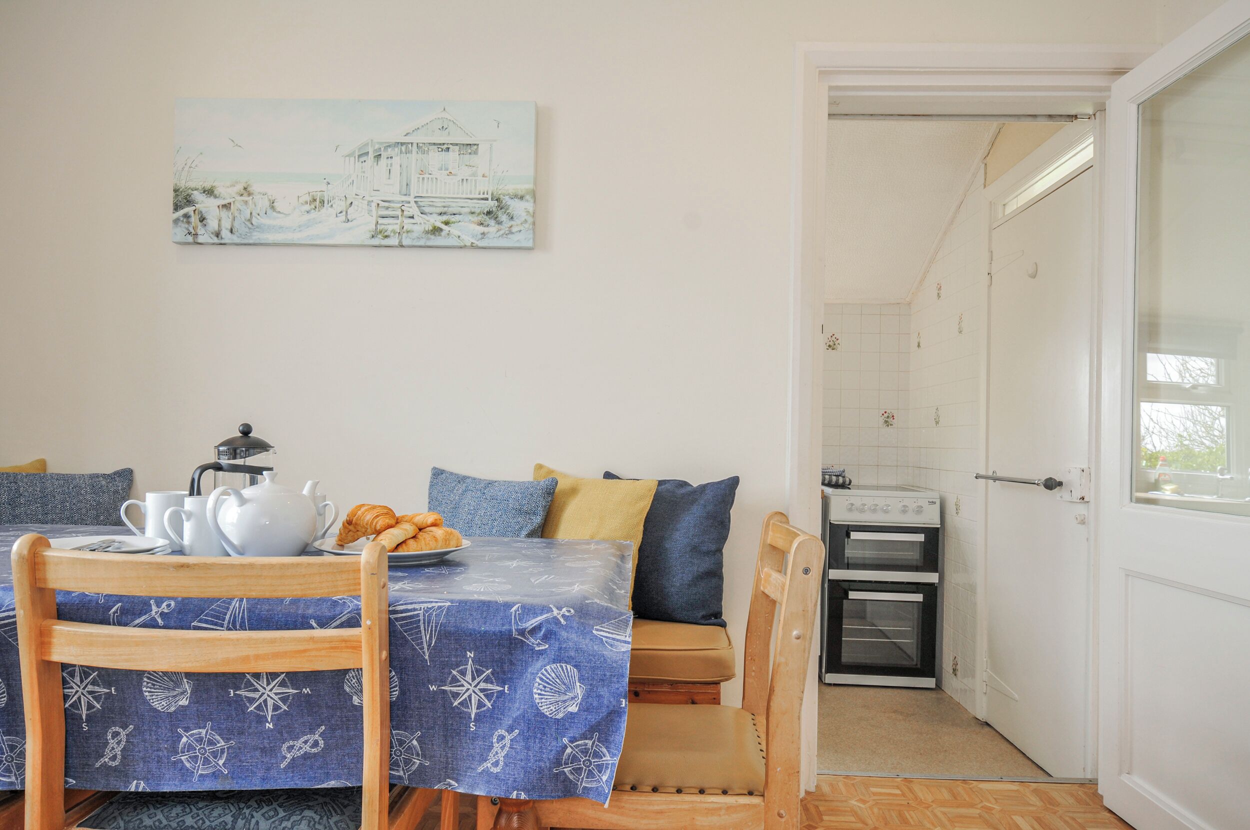Uma área de refeições com uma mesa posta para dois, um quadro acima da mesa e uma cozinha visível através de uma porta aberta.