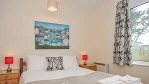 A hotel room with a bed, two bedside tables with lamps, a painting on the wall, and a window with curtains. - Hillcroft Bungalow (Wadebridge)