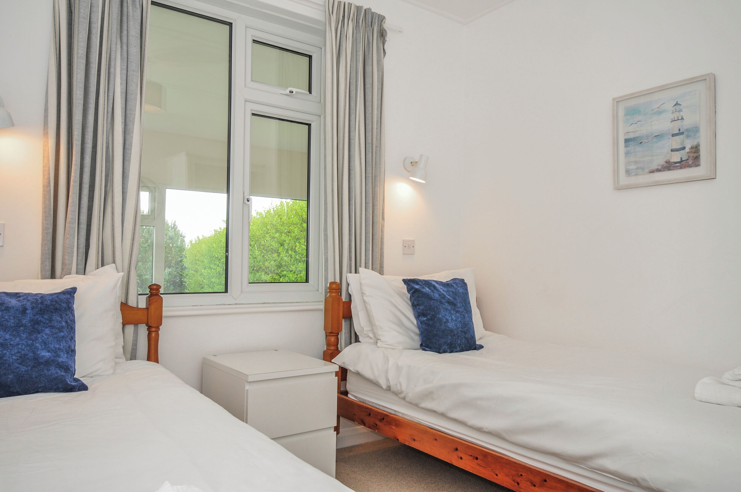A bedroom with two beds, a nightstand, a window with curtains, and a wall-mounted light.