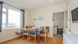 A dining area with a table set for four, a bench, and a kitchen visible through an open door. - Hillcroft Bungalow (Wadebridge)