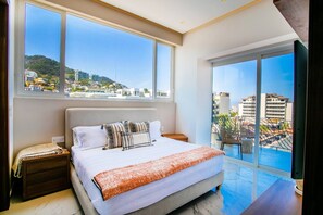 Ein Schlafzimmer mit einem großen Bett, einem Nachttisch, einem Fenster mit Stadtblick und einem Balkon mit Geländer.