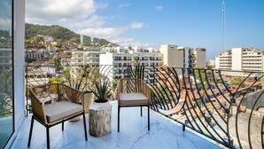 Un balcón con dos sillas de mimbre y una planta en maceta, con vistas a un paisaje urbano que incluye edificios y una colina.