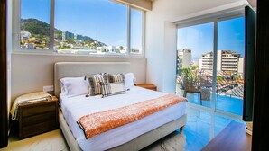 Un dormitorio con una cama grande, una mesita de noche y vistas a un paisaje urbano a través de amplias ventanas.