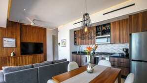 A modern kitchen with wooden cabinets, a stainless steel refrigerator, and a dining area with a wooden table and grey sofas. - Luxury D’esire 1BR w/ City View & Private BBQ (Puerto Vallarta)