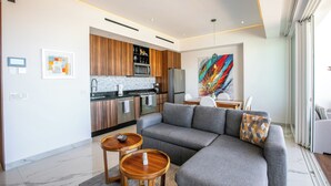 A modern living room with a grey sofa, wooden side tables, a kitchen area with wooden cabinets, and a colorful abstract painting. - Luxury D’esire 1BR w/ City View & Private BBQ (Puerto Vallarta)