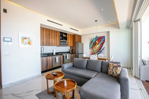 A modern living room with a grey sofa, wooden side tables, a kitchen area with wooden cabinets, and a colorful abstract painting.