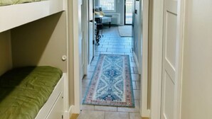 Um corredor estreito com beliches de um lado e uma porta que dá para um quarto com uma cama e uma janela.