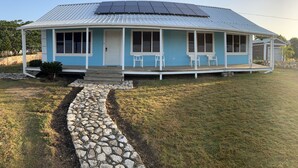 Een gelijkvloers huis met een blauwe gevel, een overdekte veranda met twee stoelen, en een pad van stenen dat naar de ingang leidt.