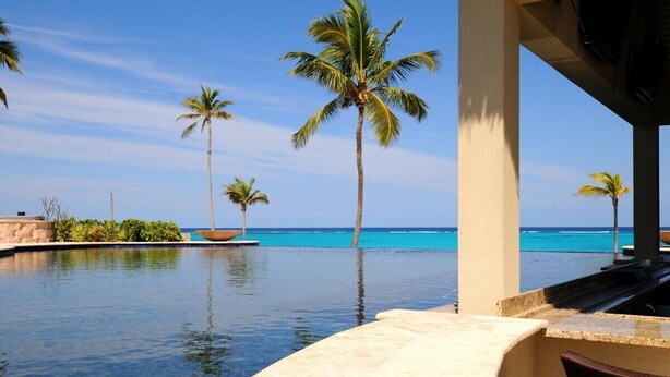 A clear view of a pool with palm trees and a beach in the background.