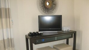 A hotel room with a flat-screen TV on a dark wooden console table, two beige ottomans, and a decorative wall piece above the TV.