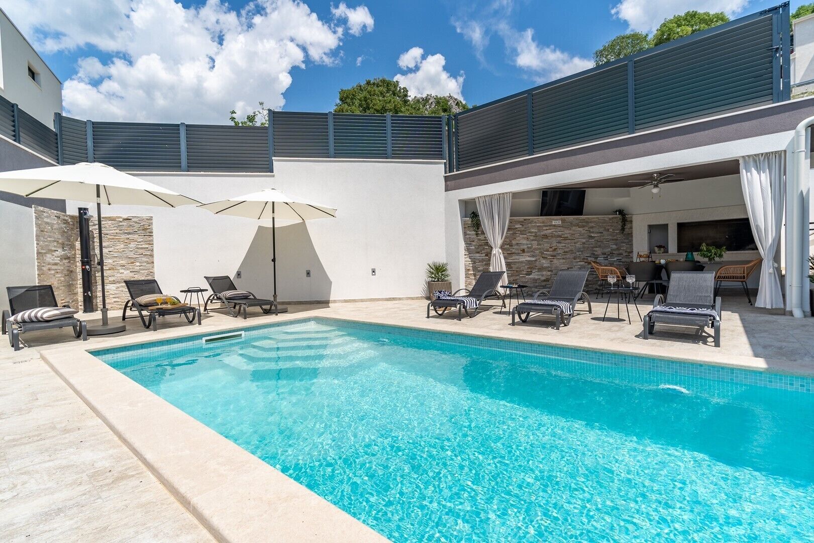 A swimming pool with lounge chairs and umbrellas, a covered patio area, and a stone wall.