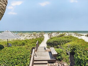 A wooden pathway leading to a beach with a striped umbrella and benches.